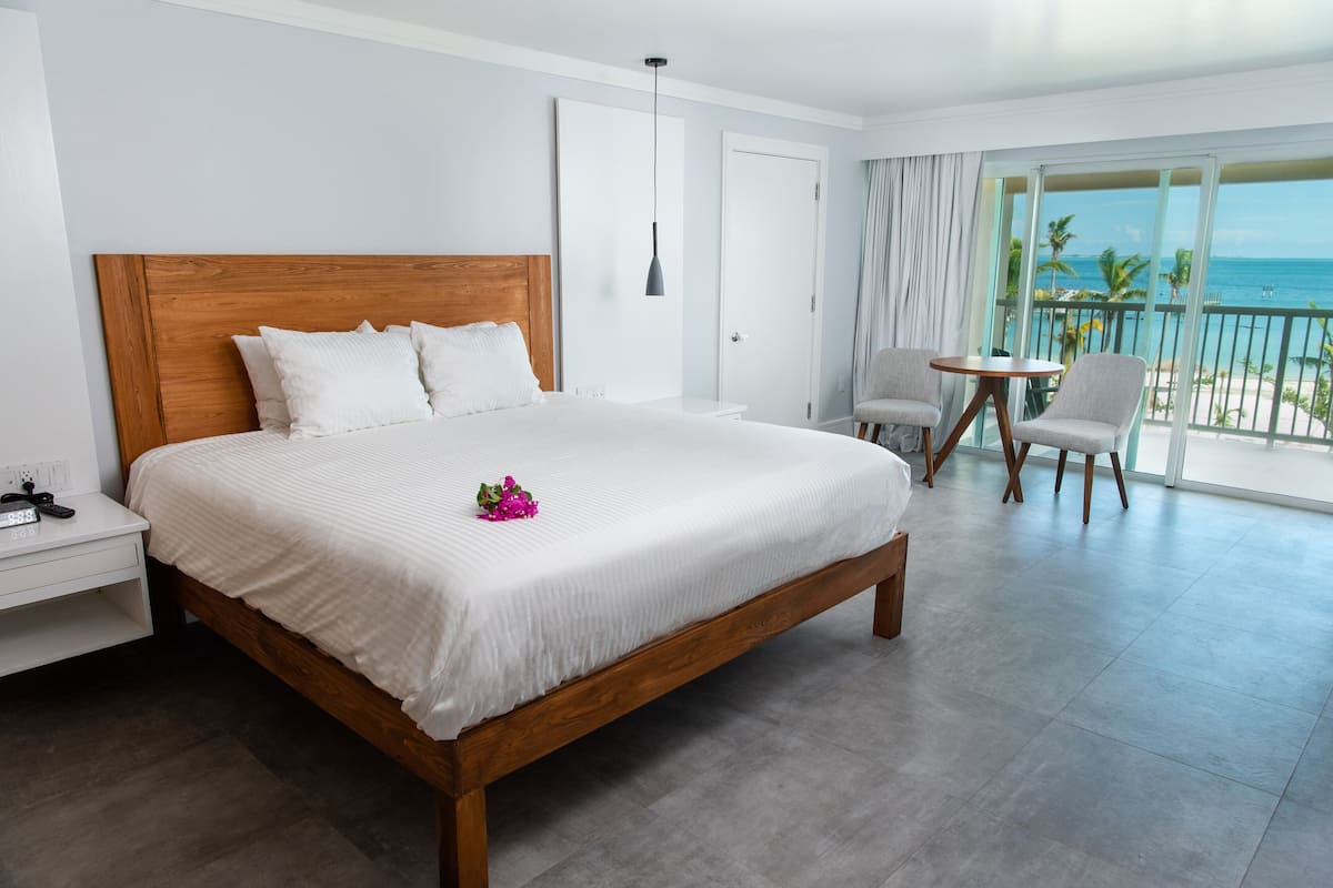 Una cama grande con cabecera de madera, una mesita de noche con teléfono y cargador, una pequeña mesa redonda con dos sillas y un balcón con vista al océano.