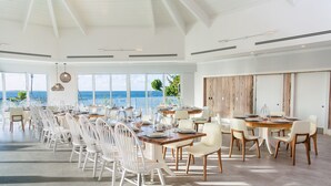 Un vaste espace dédié à la salle à manger, avec de nombreuses tables et chaises, de grandes fenêtres offrant une vue sur l’océan et un plafond haut avec des poutres apparentes.