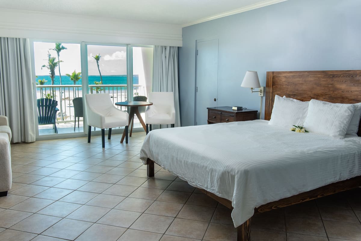 Habitación de hotel con una cama grande, una mesita, una silla y vista al océano a través de una puerta corrediza.