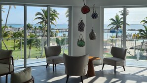 Une pièce spacieuse avec de grandes fenêtres donnant sur un port de plaisance et des palmiers. On y trouve quatre chaises et une petite table.