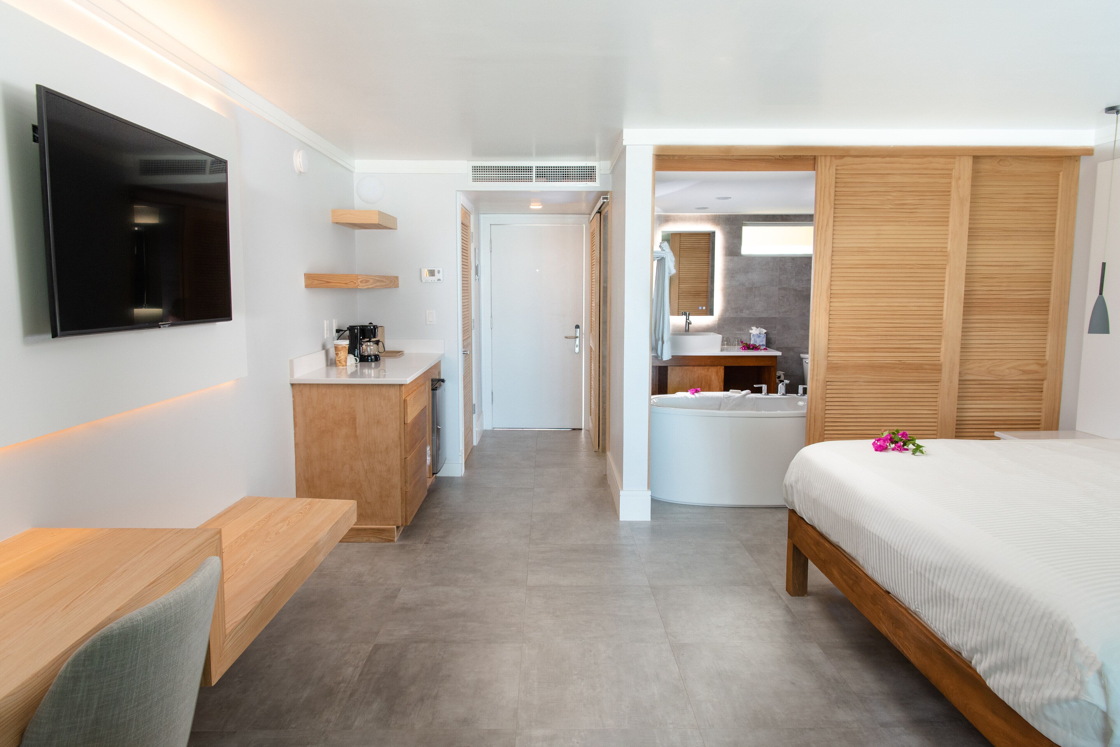 A modern hotel room with a flat-screen TV, wooden furniture, a large bed, a bathroom with a bathtub, and a walk-in closet.