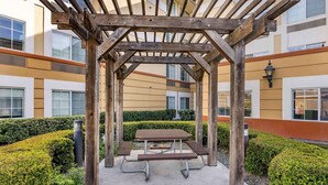 A wooden pergola with a picnic table in an outdoor courtyard surrounded by hedges and a building with large windows.