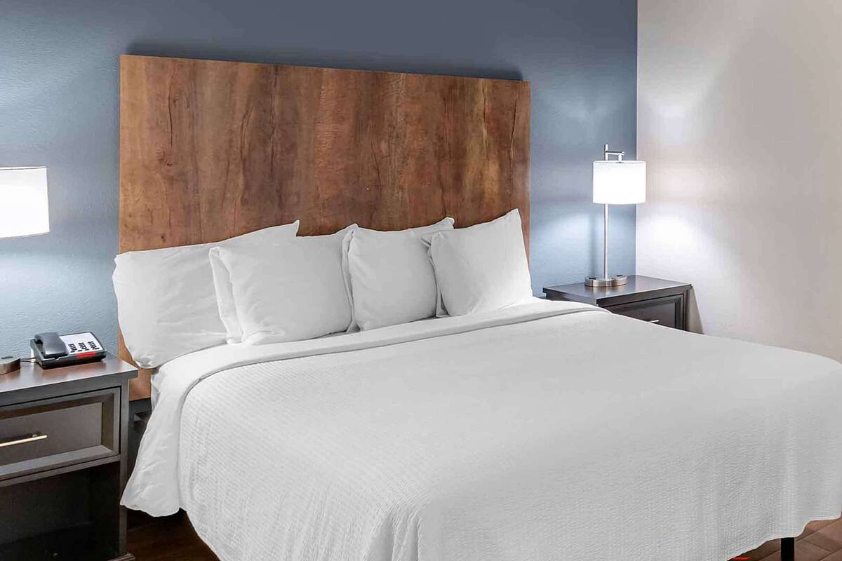 Een hotelkamer met een groot bed, een houten hoofdbord, witte beddengoed, een nachtkastje met een lamp en een telefoon.