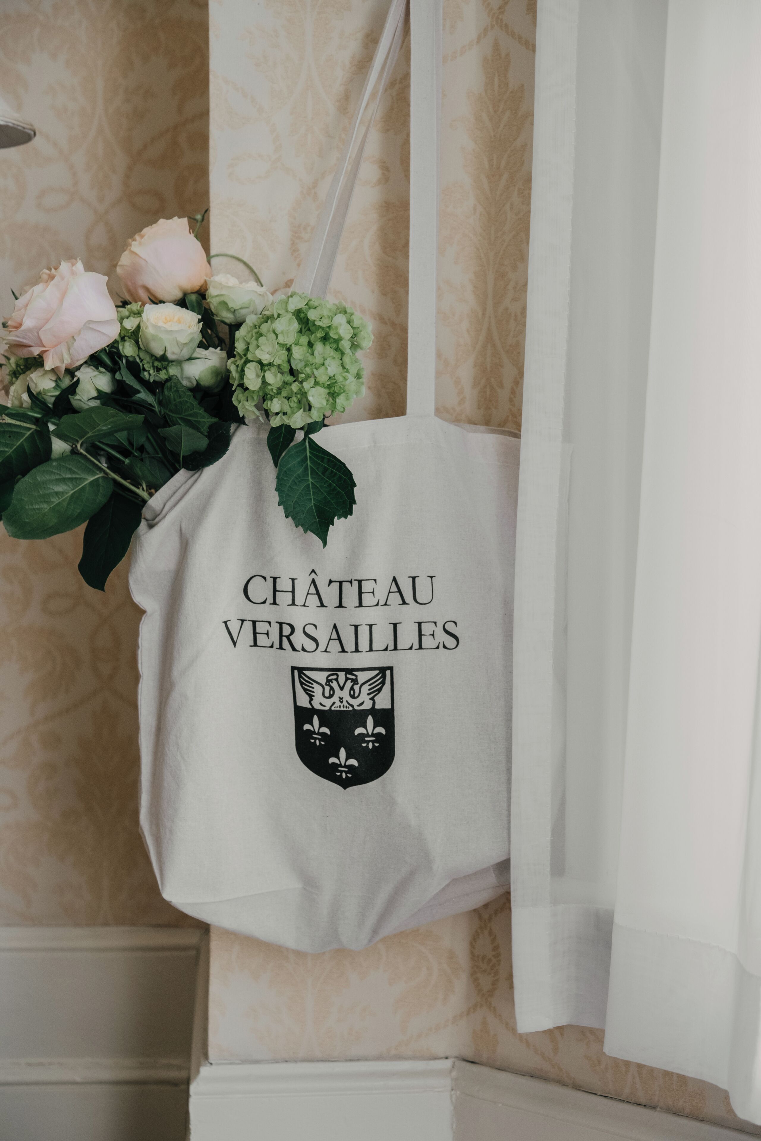 A white tote bag with 'CHÂTEAU VERSAILLES' and a crest logo, hanging on a wall with patterned wallpaper.
