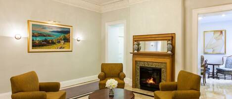 A formal sitting room with a fireplace, four armchairs, a round table with a vase, and two paintings.