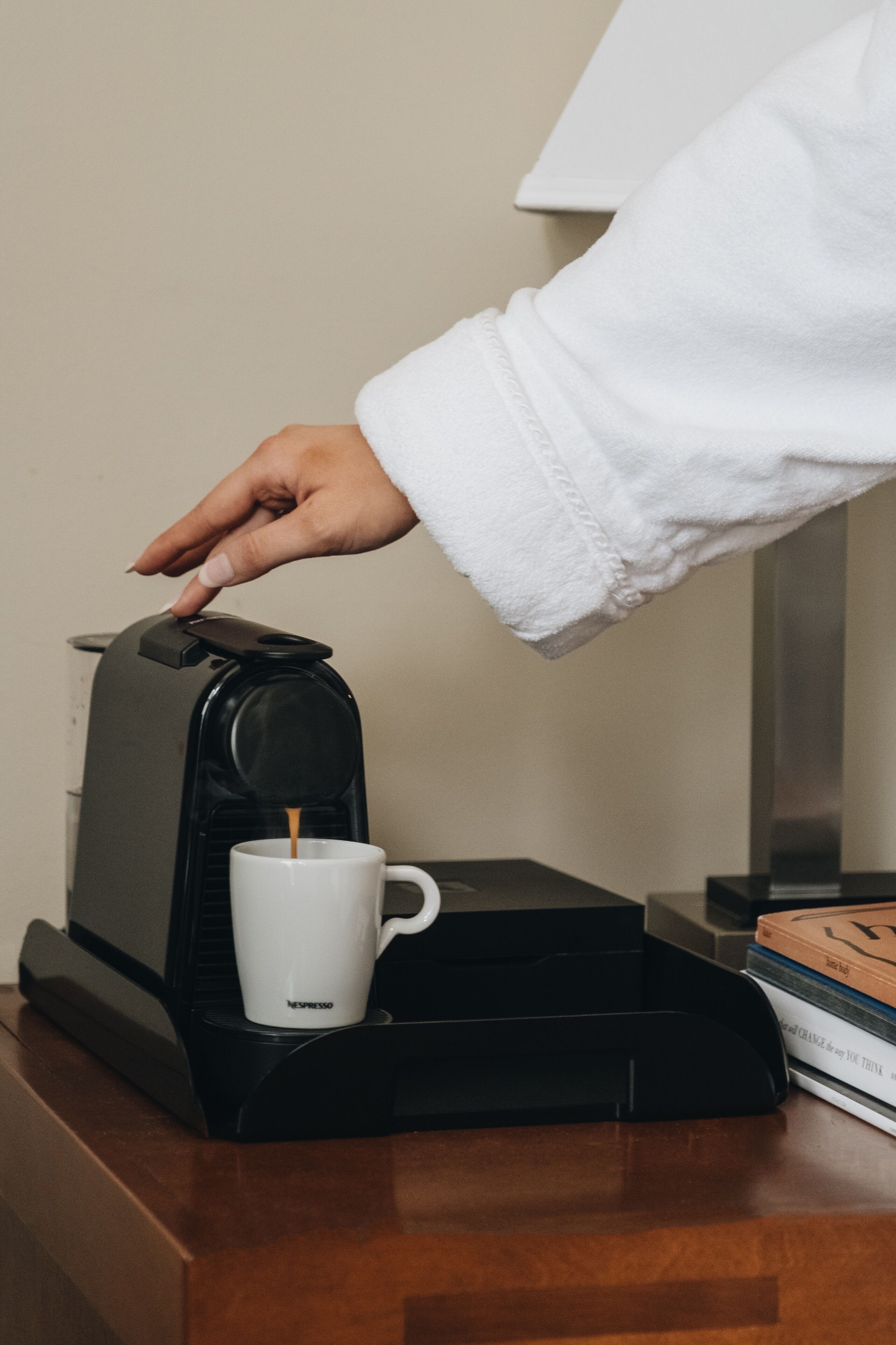 A hand pressing a button on a coffee machine, a white cup, and a book on a wooden surface.