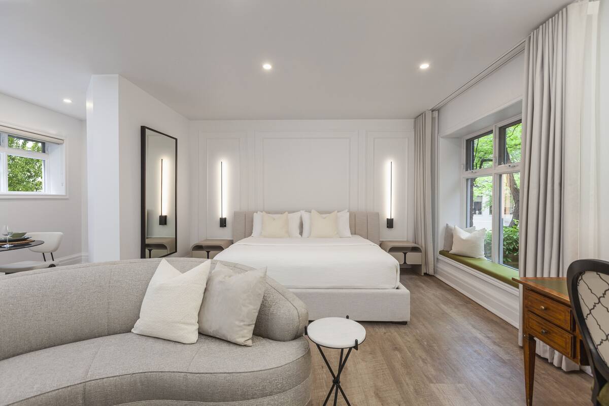 A modern bedroom with a large bed, a sofa, a small table, and a window with a view of greenery.