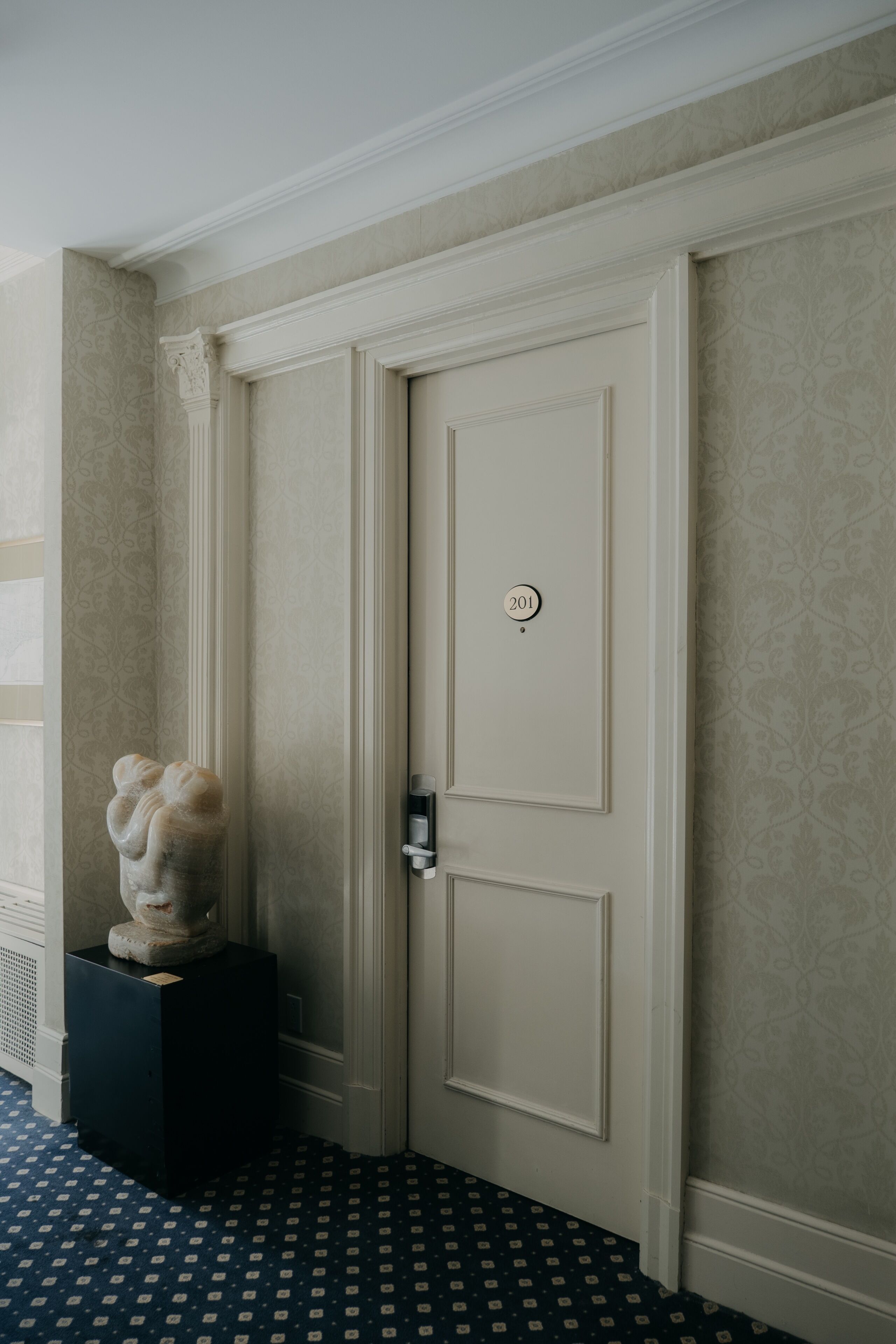 A room with a closed door marked '201,' a marble bust on a pedestal, patterned wallpaper, and a patterned floor.