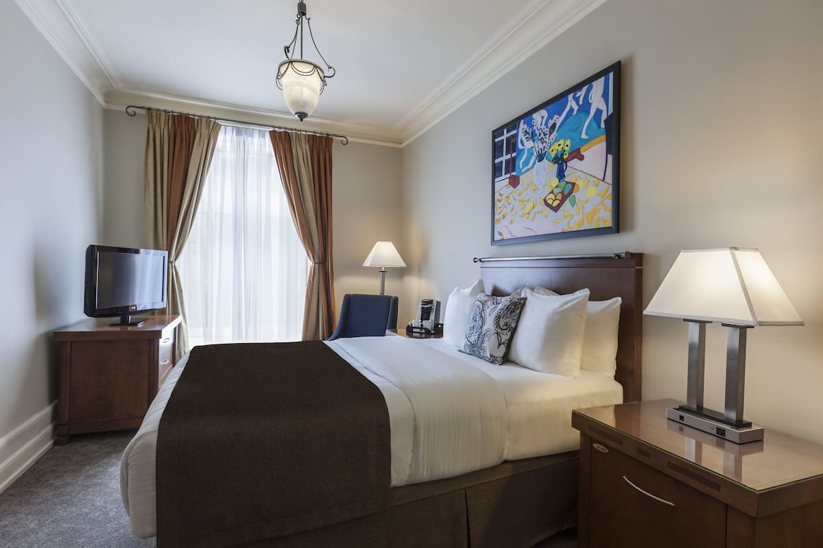 A hotel room with a bed, a TV, a desk with a lamp, and a painting on the wall.