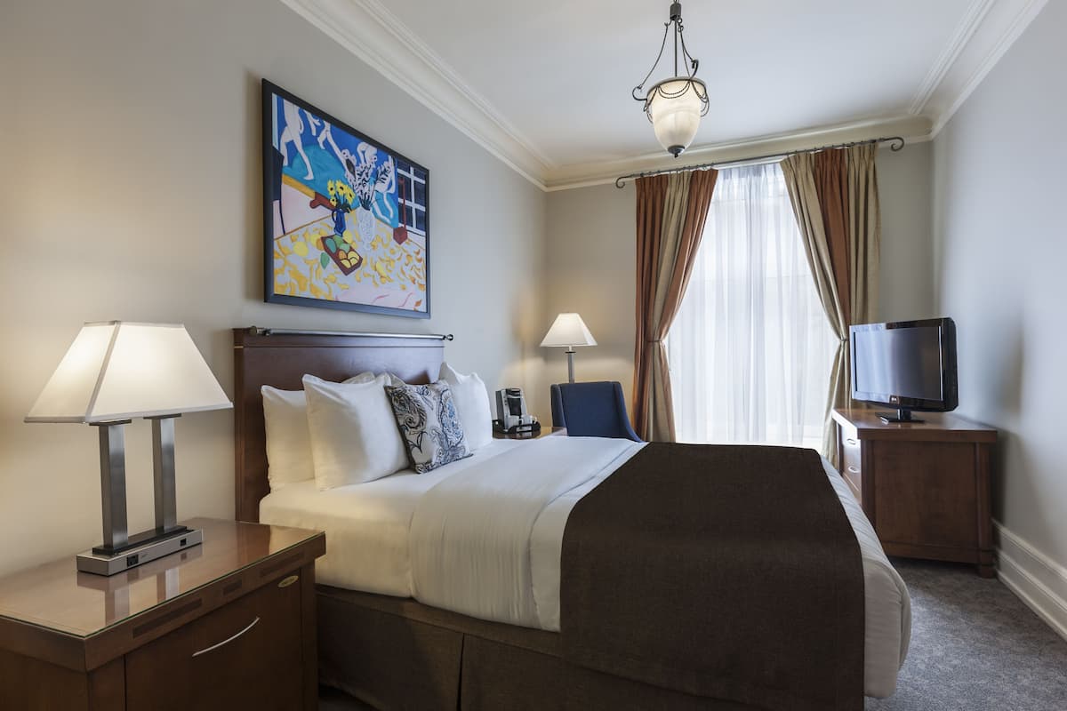 A hotel room with a large bed, two bedside lamps, a flat-screen TV, a painting on the wall, and a window with curtains.