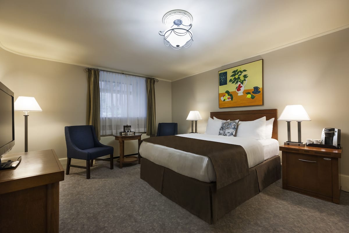 A hotel room with a bed, two bedside tables with lamps, a desk with a computer, a chair, a small table with a lamp, and a painting on the wall.