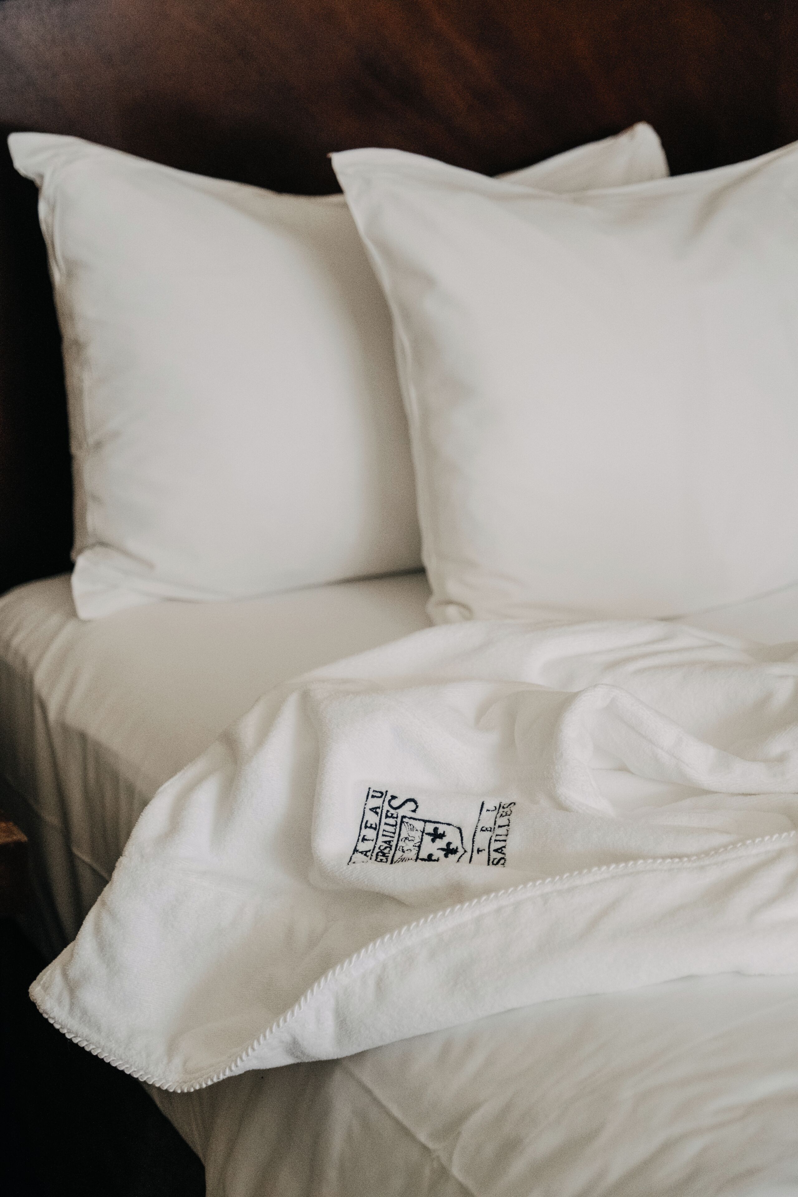 A bed with white bedding and pillows, a visible logo on the blanket, and a wooden headboard.