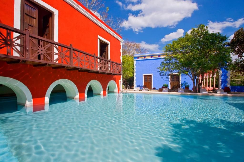 Una piscina con agua azul cristalina, un edificio rojo con arcos blancos y un edificio azul con una puerta.