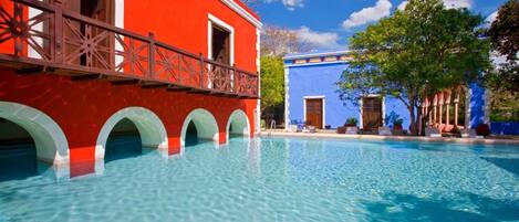 A swimming pool with clear blue water, a red building with white arches, and a blue building with a door.