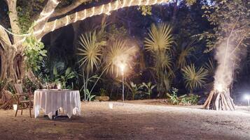 Un espacio de comedor al aire libre con una mesa dispuesta para una comida, rodeado de plantas tropicales y un árbol grande.