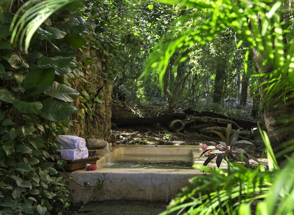 A serene garden with a small pond, lush greenery, and a stone bench with a basket and a rolled-up mat.