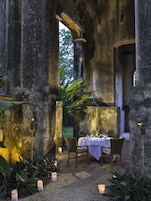 Un área de comedor al aire libre con una mesa dispuesta para dos personas, rodeada de columnas de piedra y plantas.