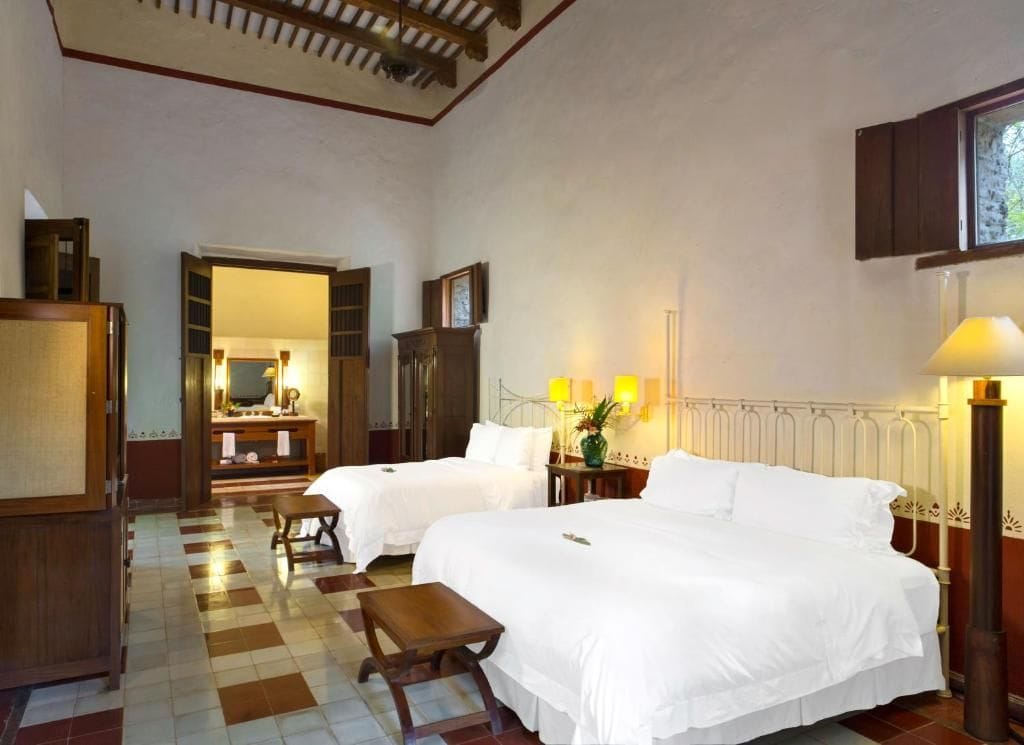 Habitación de hotel con dos camas, un banco de madera, un escritorio con lámpara y vistas al exterior por una ventana.