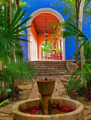A stone fountain with water and red flowers, surrounded by greenery and a blue archway leading to a seating area with chairs and a table.
