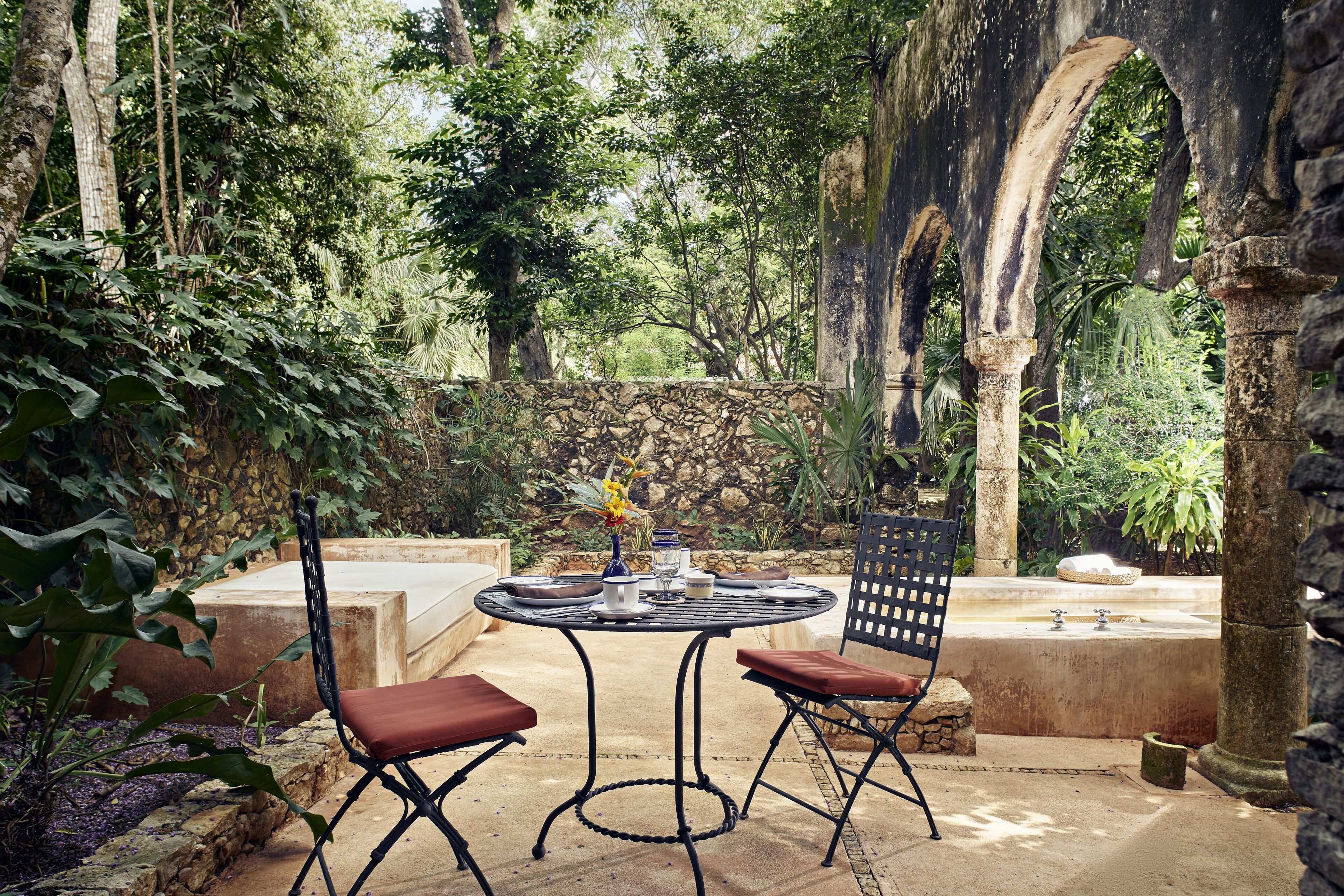 An outdoor patio with a round table and two chairs, surrounded by lush greenery and stone structures.