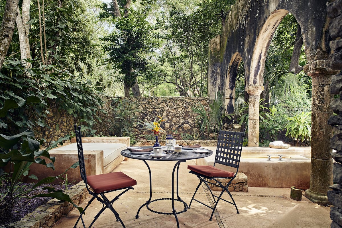 An outdoor patio with a round table and two chairs, surrounded by lush greenery and stone structures.