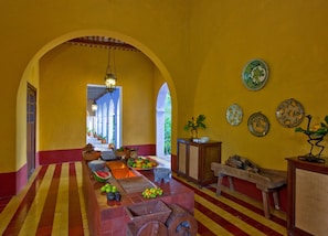 A corridor with a tiled floor, a central table with fruits and vegetables, and a wall with decorative plates.