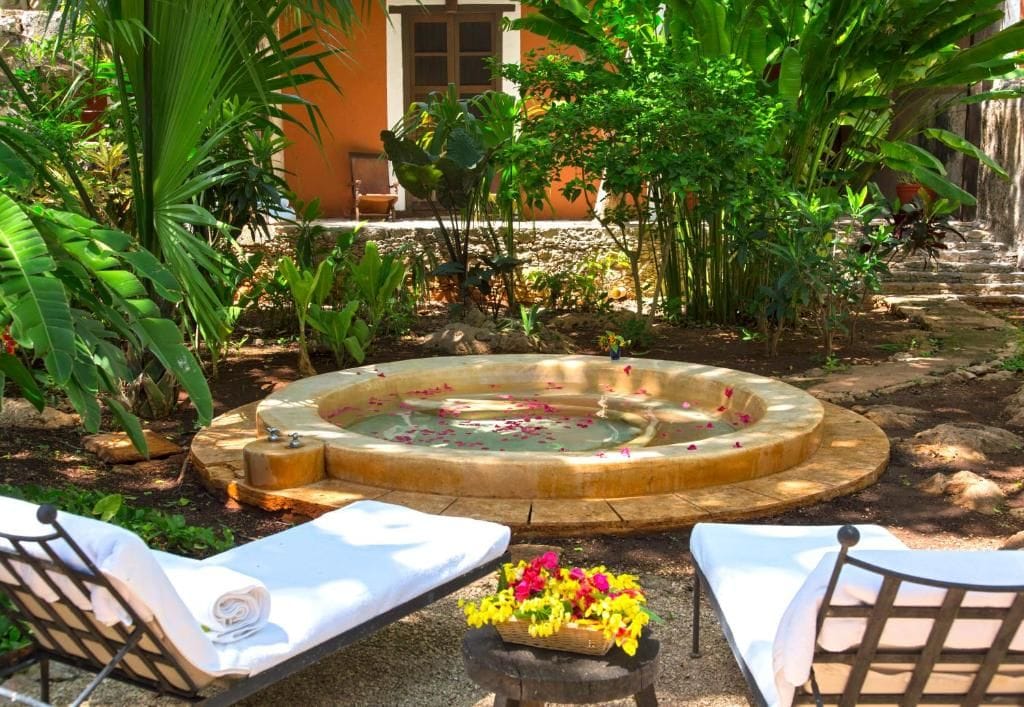 Un jacuzzi rodeado de exuberante follaje, con dos sillones de descanso y un arreglo floral en primer plano.
