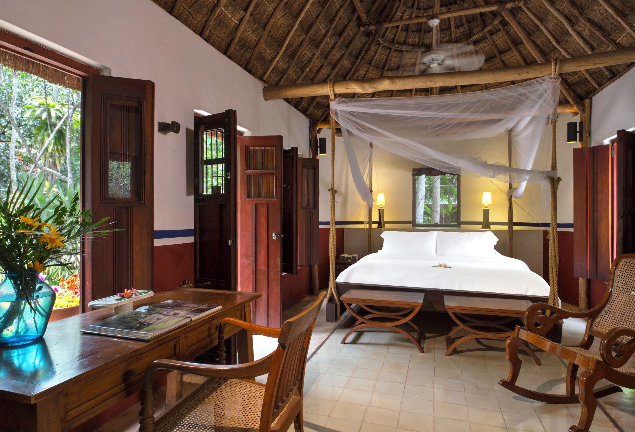 A room with a bed, wooden furniture, a table with a vase, and a window with a view of greenery.