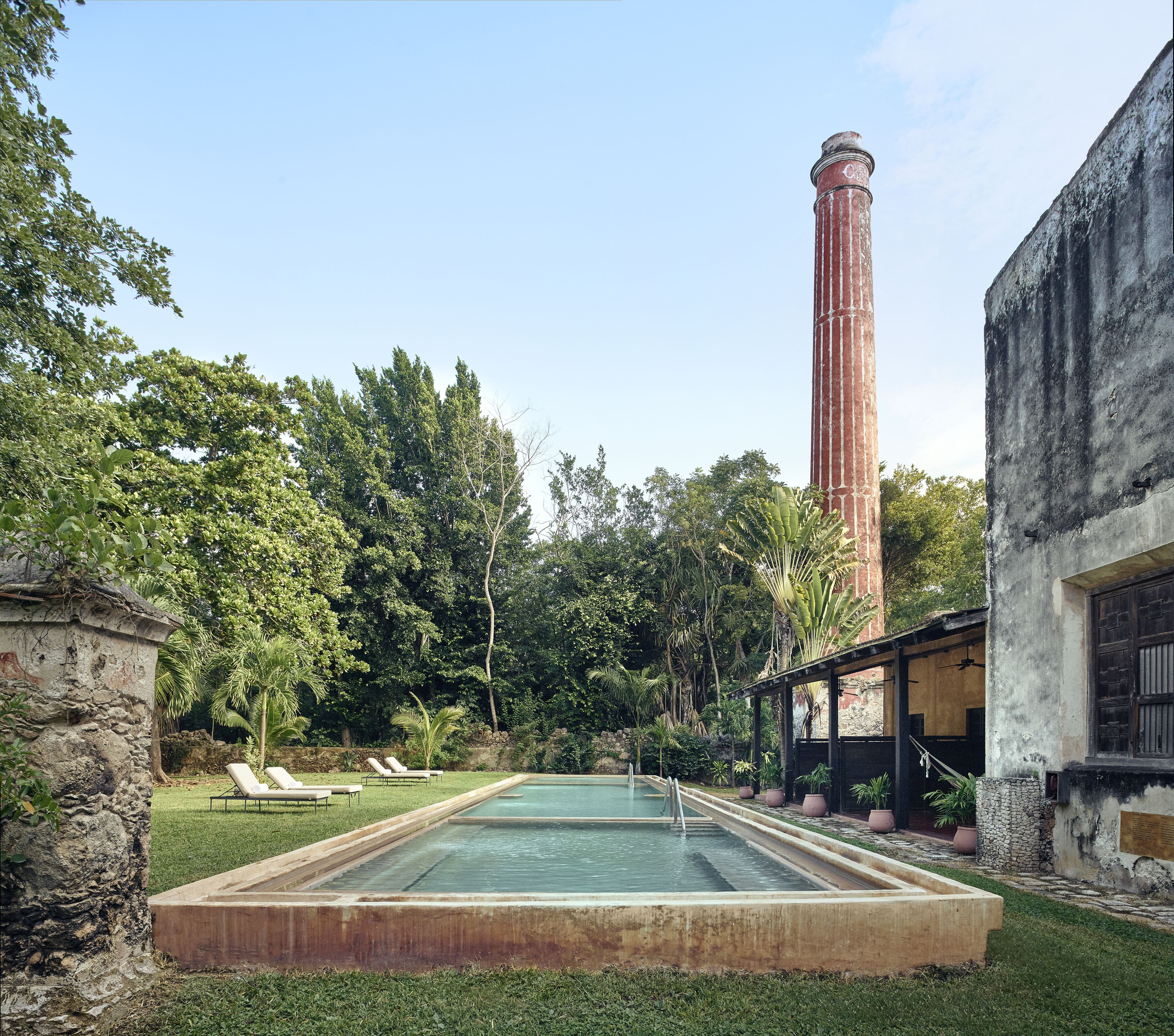 A swimming pool with a stone border, surrounded by lush greenery and a tall brick chimney.
