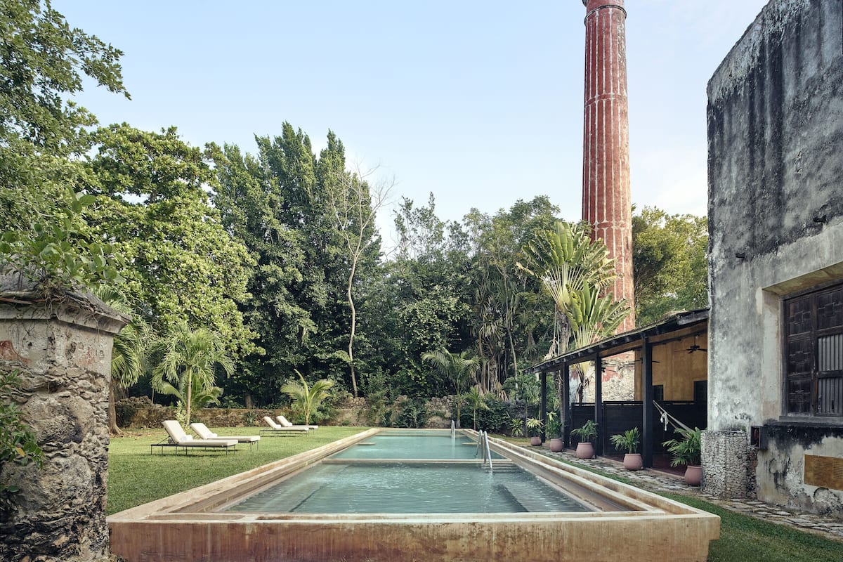 A swimming pool with a stone border, surrounded by lush greenery and a tall brick chimney.
