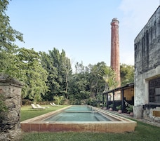 Una piscina con borde de piedra, rodeada de exuberante vegetación y una alta chimenea de ladrillo.