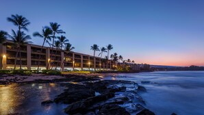 A beachfront hotel with multiple floors, palm trees, and a rocky shoreline.
