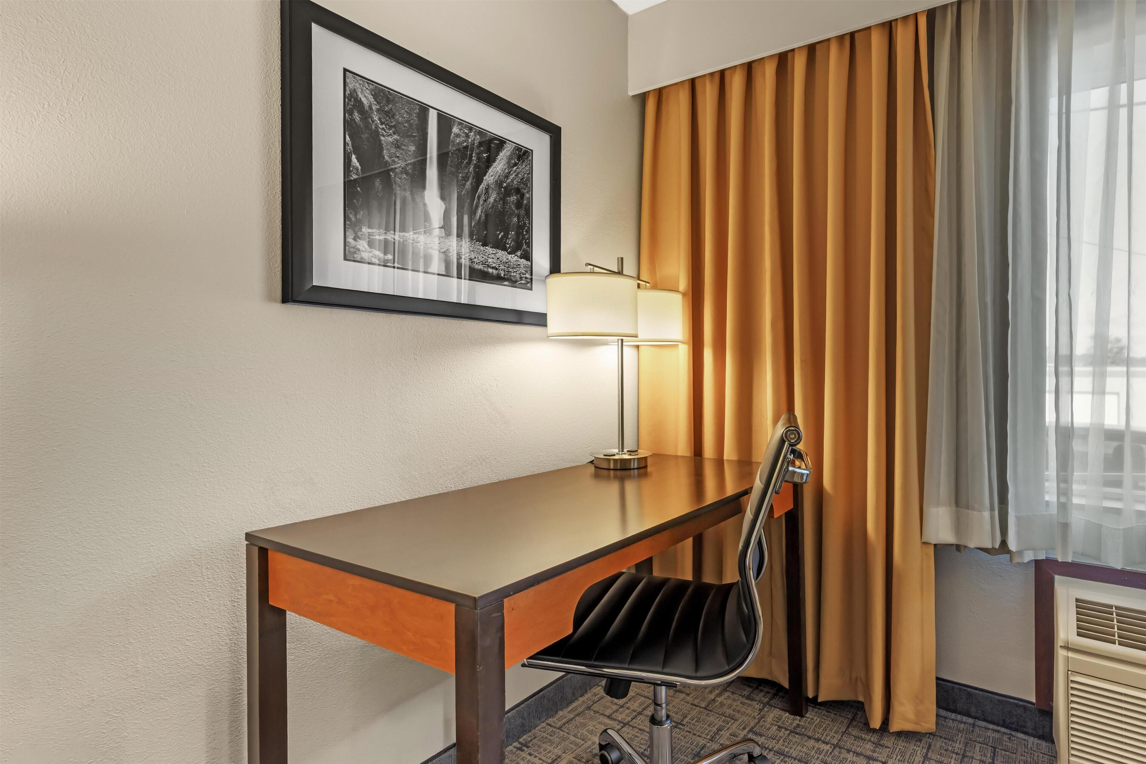 Een hotelkamer met een bureau, stoel, lamp en een ingelijste foto aan de muur.