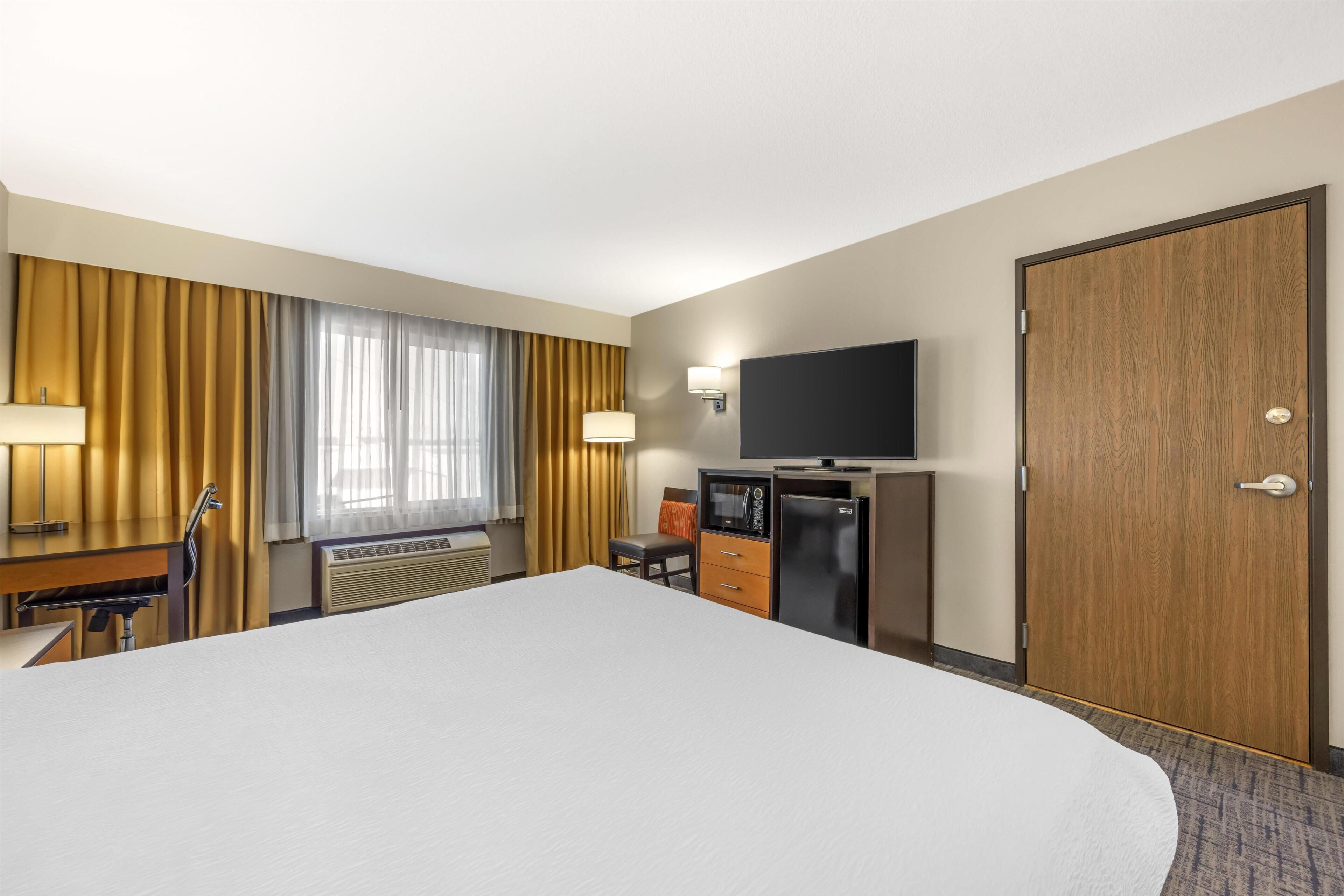 Una habitación de hotel con una cama grande, un escritorio con silla, televisión, minibar y una puerta de madera.