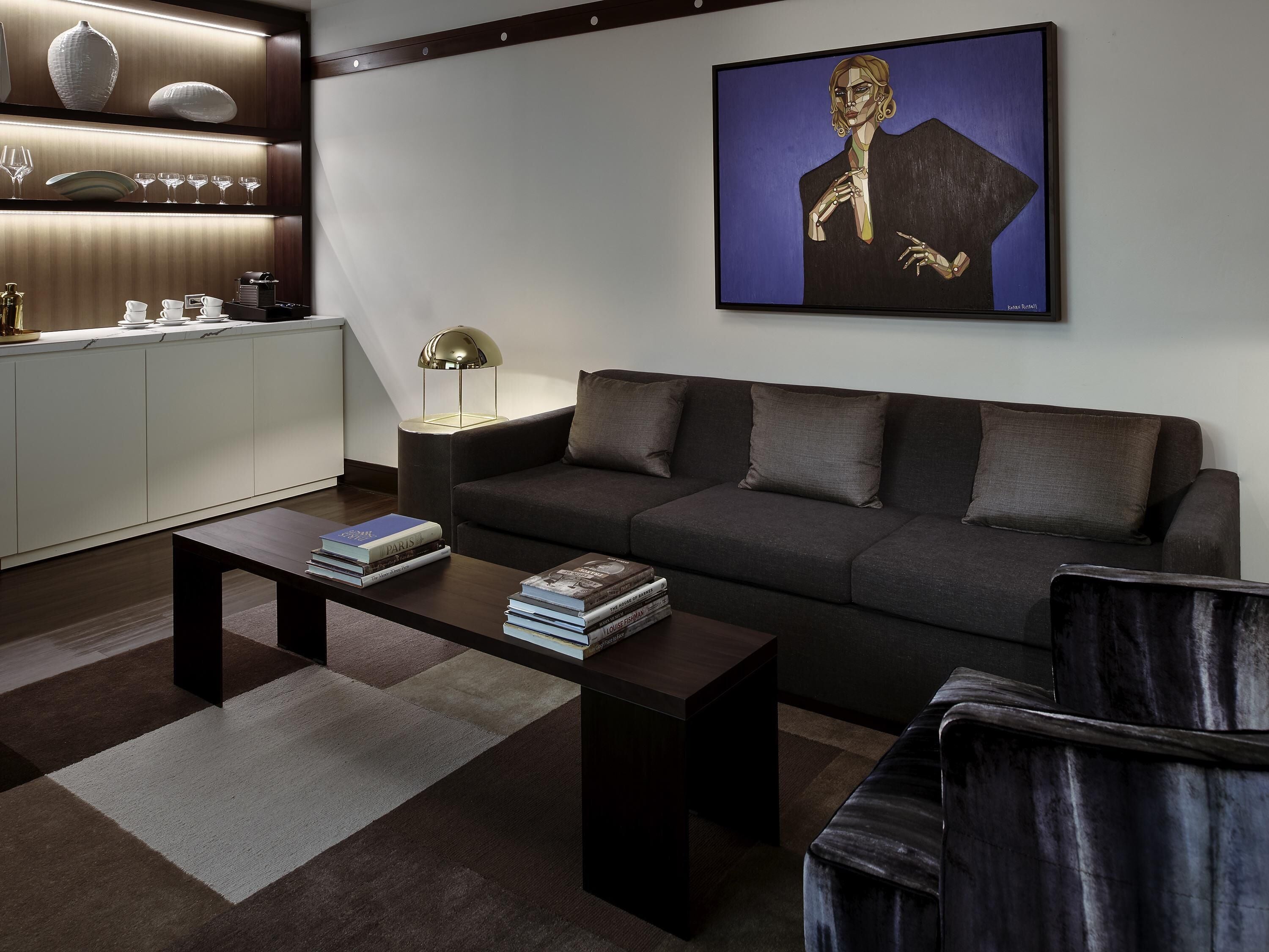 A modern living room with a sofa, coffee table, and armchair, a painting on the wall, and a kitchen area with cabinets and a lamp.