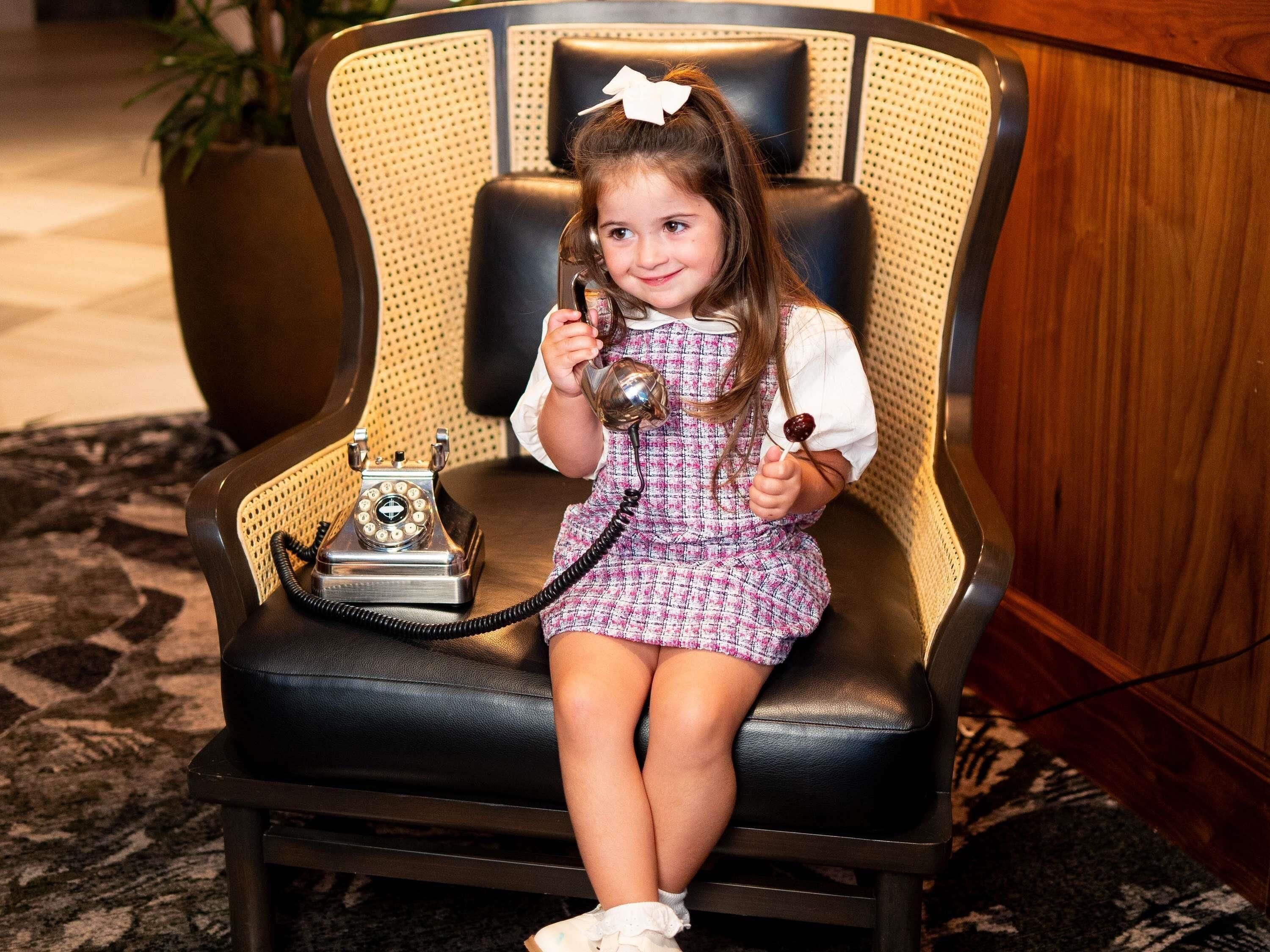 A child sitting on a vintage-style chair with a telephone.