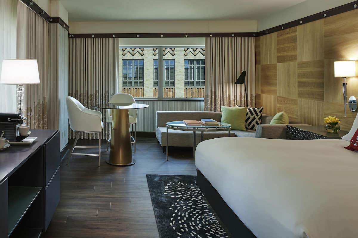 A hotel room with a bed, a desk with a lamp, a chair, a sofa, and a view of the outside.