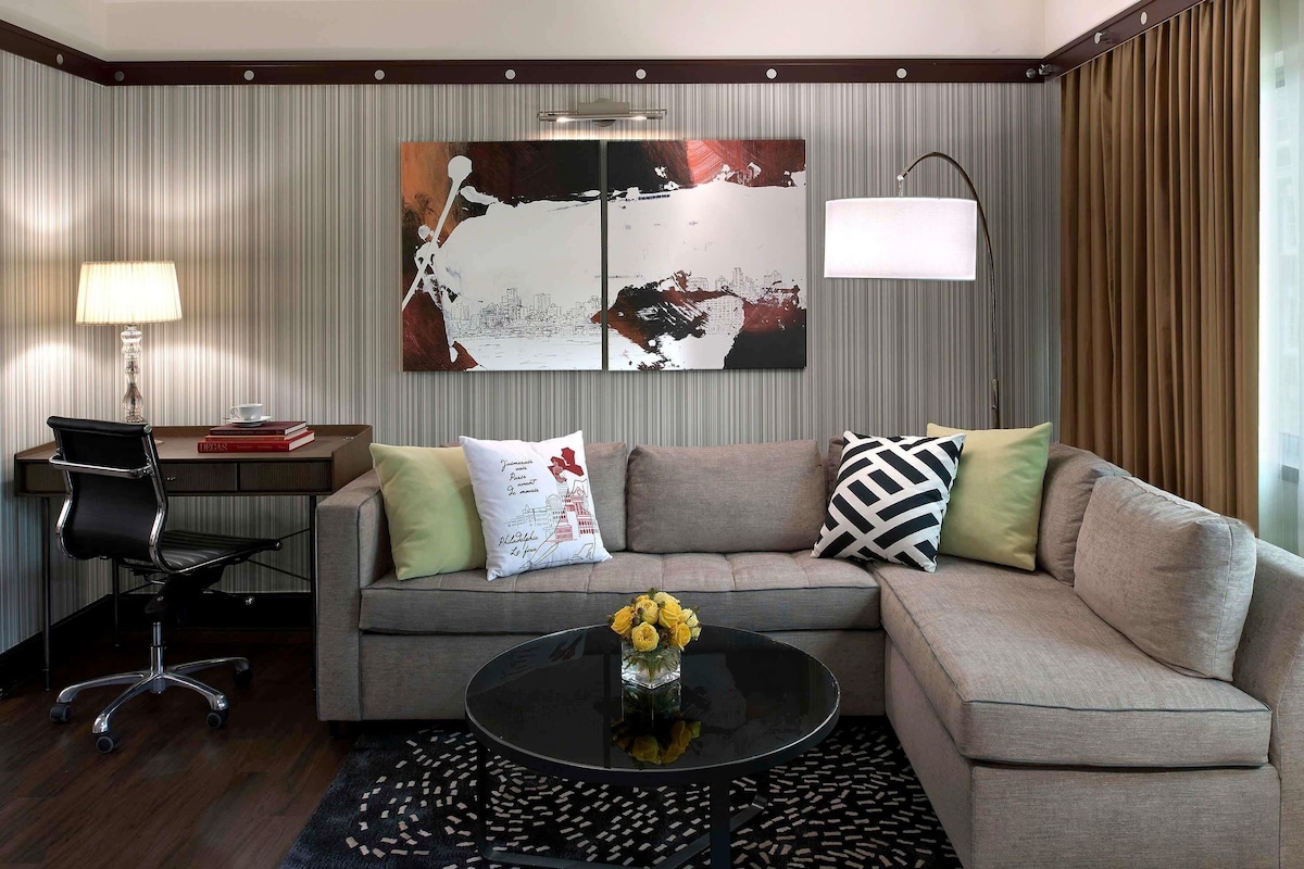 A modern living room with a sectional sofa, a glass coffee table with flowers, a desk with a lamp, and a large abstract painting on the wall.