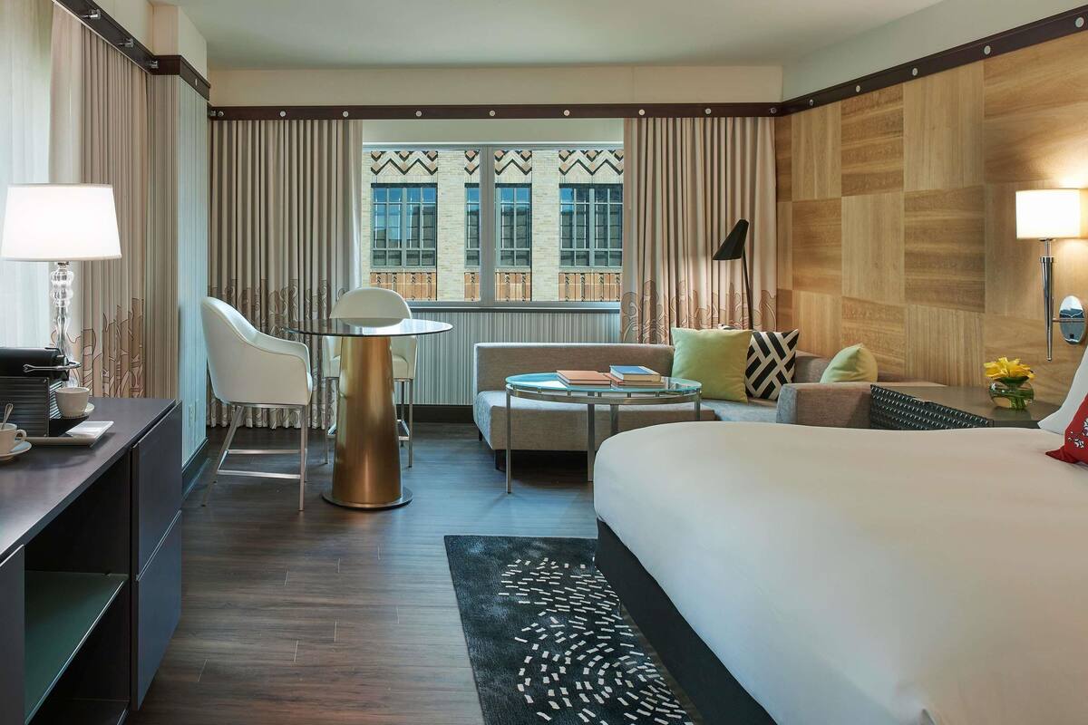 Habitación de hotel con cama, escritorio con lámpara, silla, sofá, pequeña mesa y vistas al exterior a través de amplios ventanales.