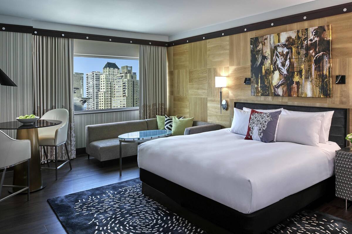 Habitación de hotel con cama, sofá, mesa de comedor y vistas a edificios altos.