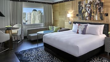 A hotel room with a bed, a sofa, a dining table, and a view of high-rise buildings.