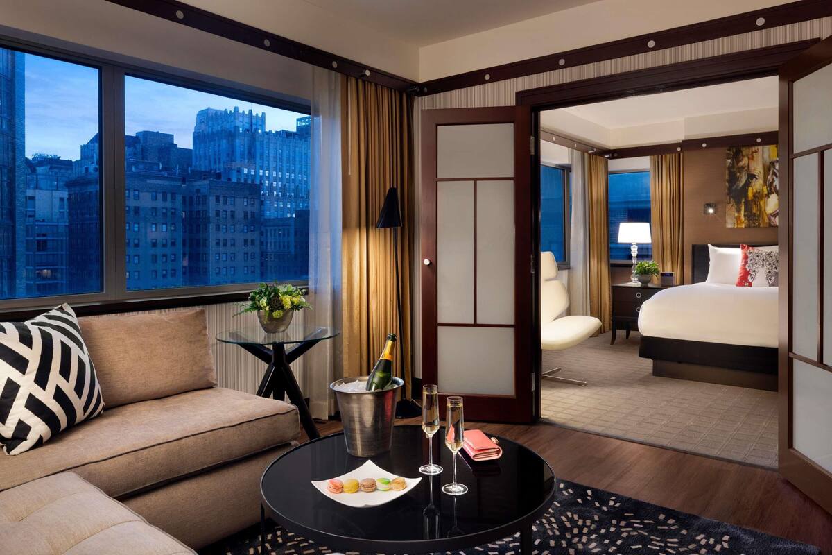 Habitación de hotel con sofá, una mesa de comedor y una botella de champán, y vista al paisaje urbano.