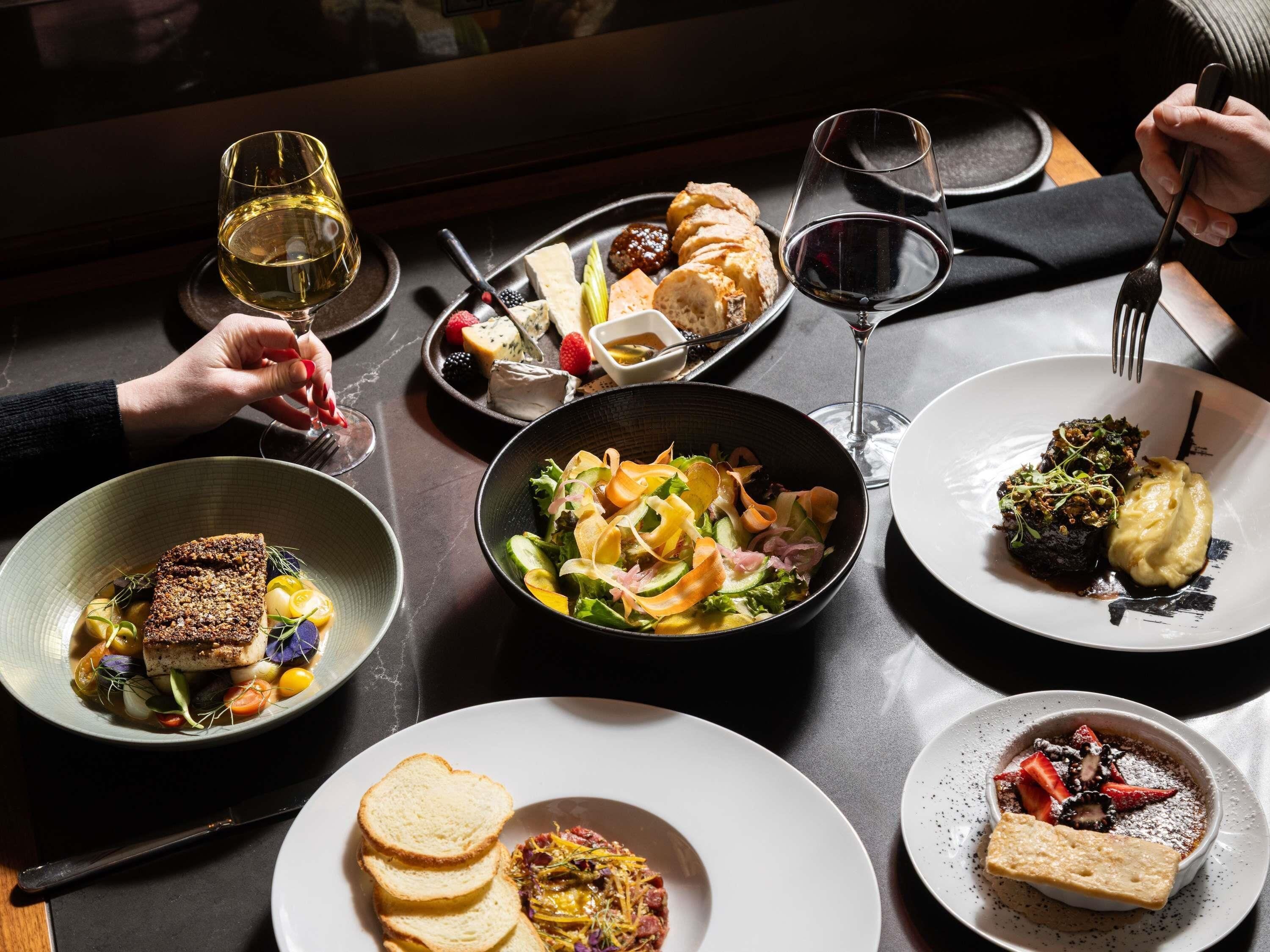 A table set with various dishes, including a fish dish, salad, and dessert, with wine glasses and cutlery.
