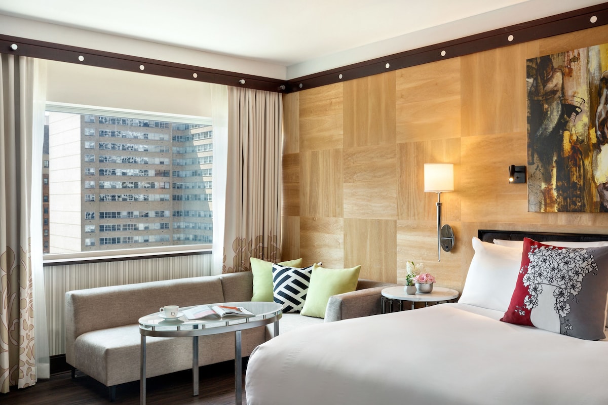 A hotel room with a large bed, a sofa, a small table, and a view of a cityscape through a window.