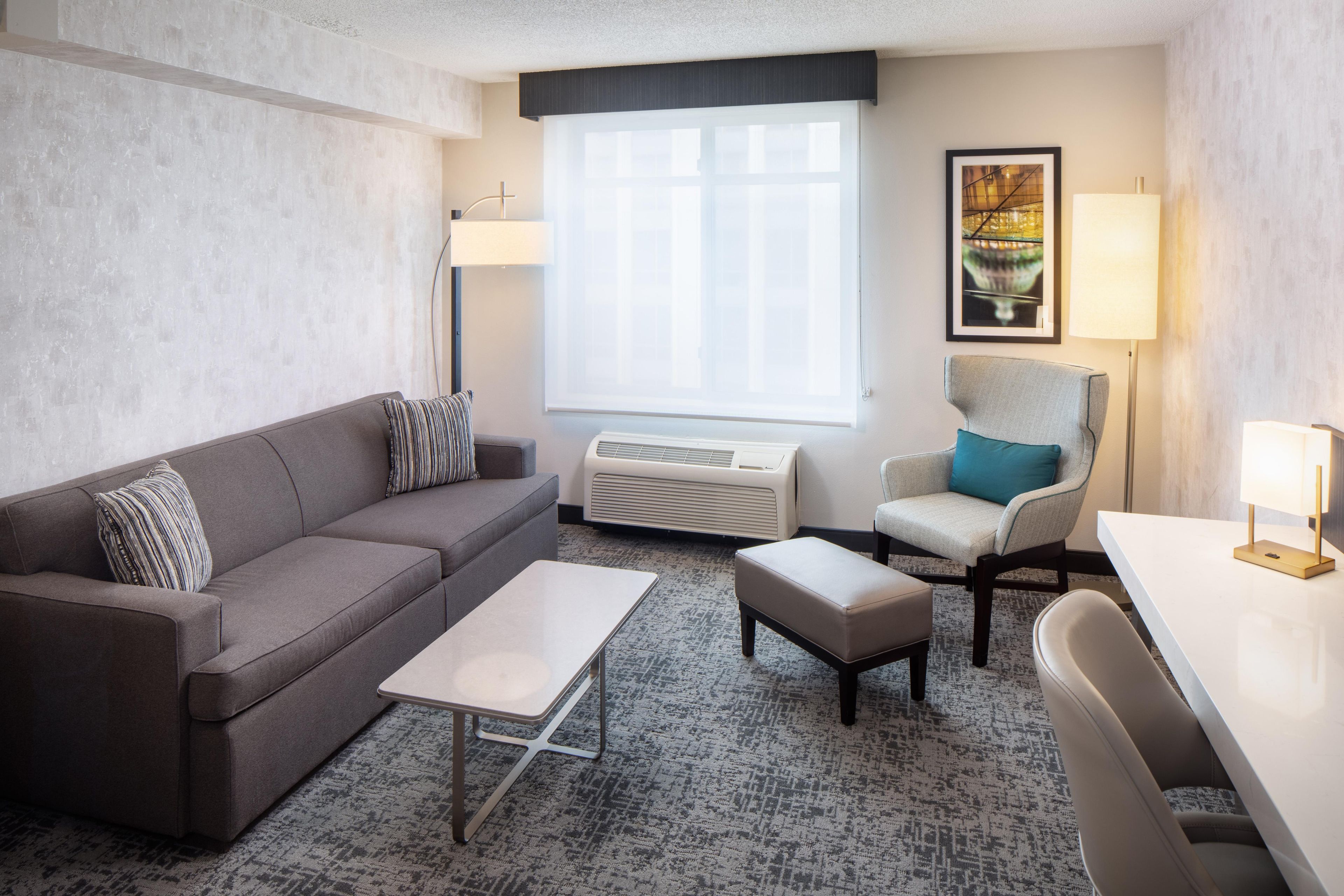 Una habitación de hotel moderna con un sofá gris, una mesita blanca pequeña, un sillón gris y un ventanal grande con persianas.