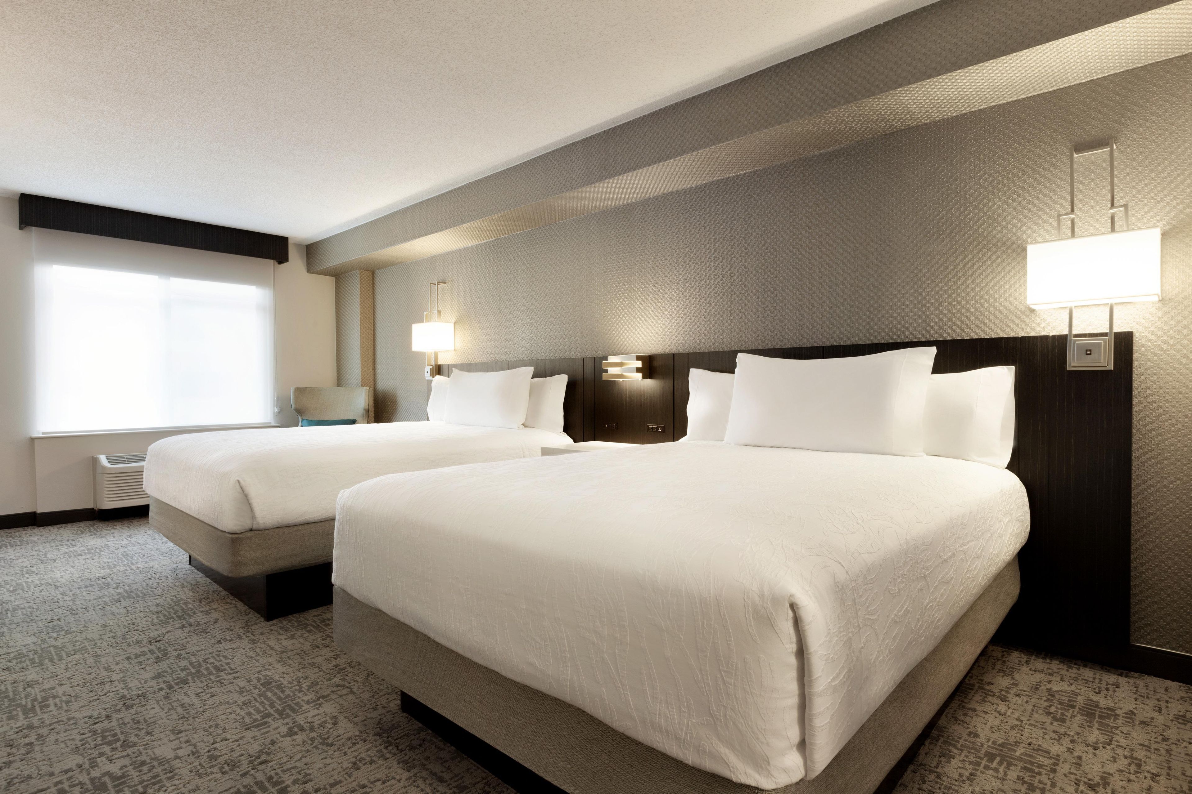 A hotel room with two beds, a large window, and a wall-mounted lamp.