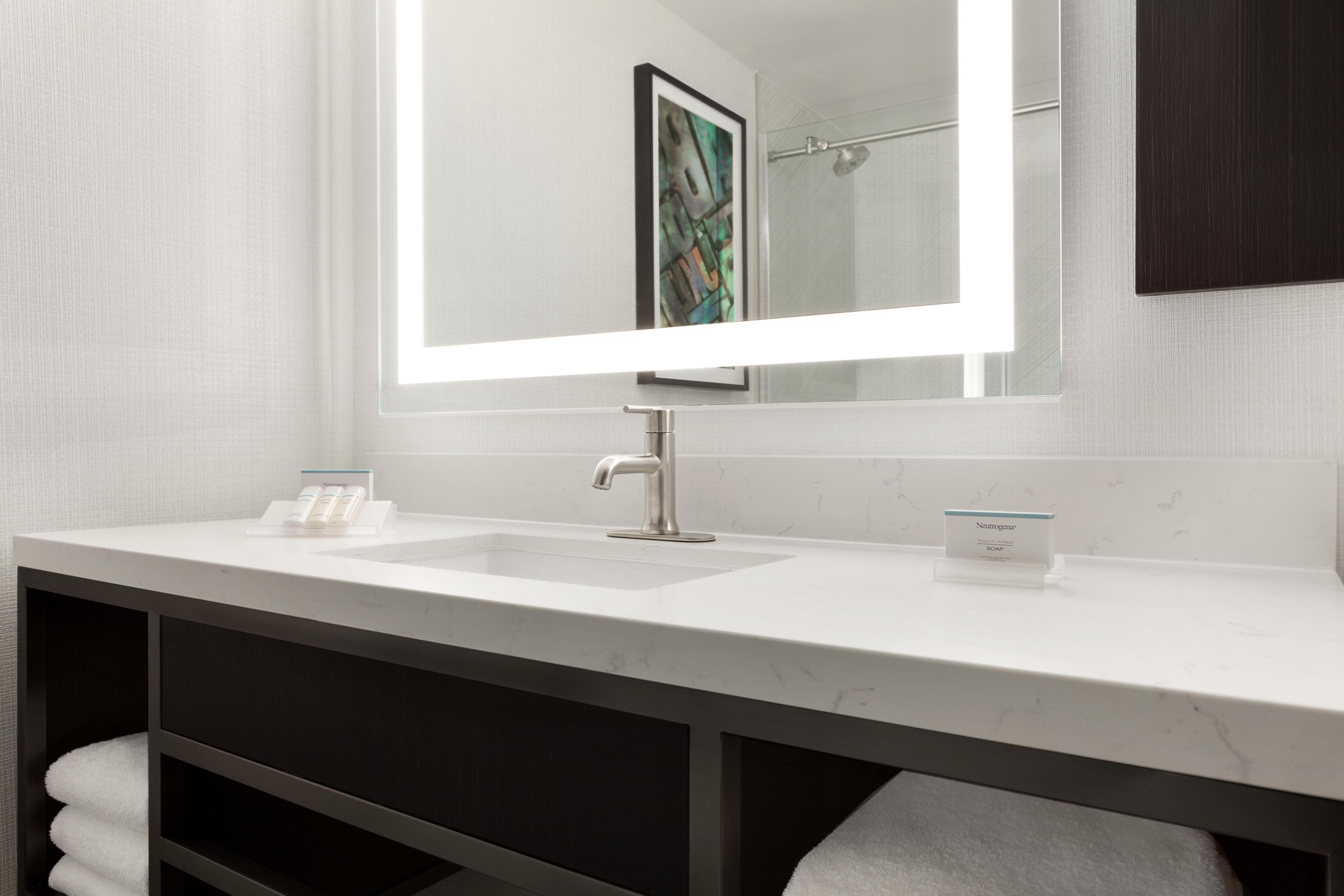 Un baño con un lavabo blanco, un espejo grande y una obra de arte enmarcada sobre el espejo.