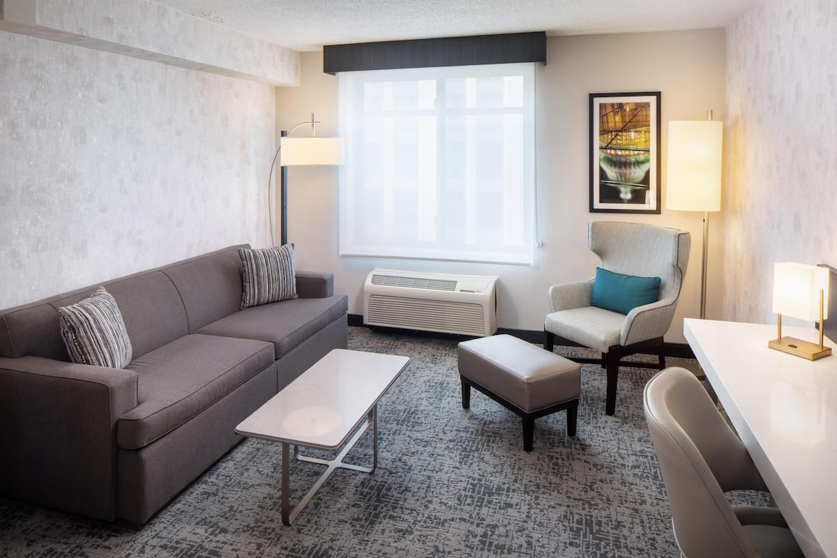 A hotel room with a gray sofa, a small table, a chair, an air conditioning unit, and a window with a view.