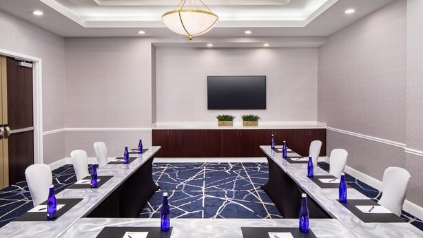 A conference room with a long table, white chairs, blue water bottles, a flat-screen TV, and a patterned carpet. - Hilton Garden Inn Washington DC Downtown (Washington)