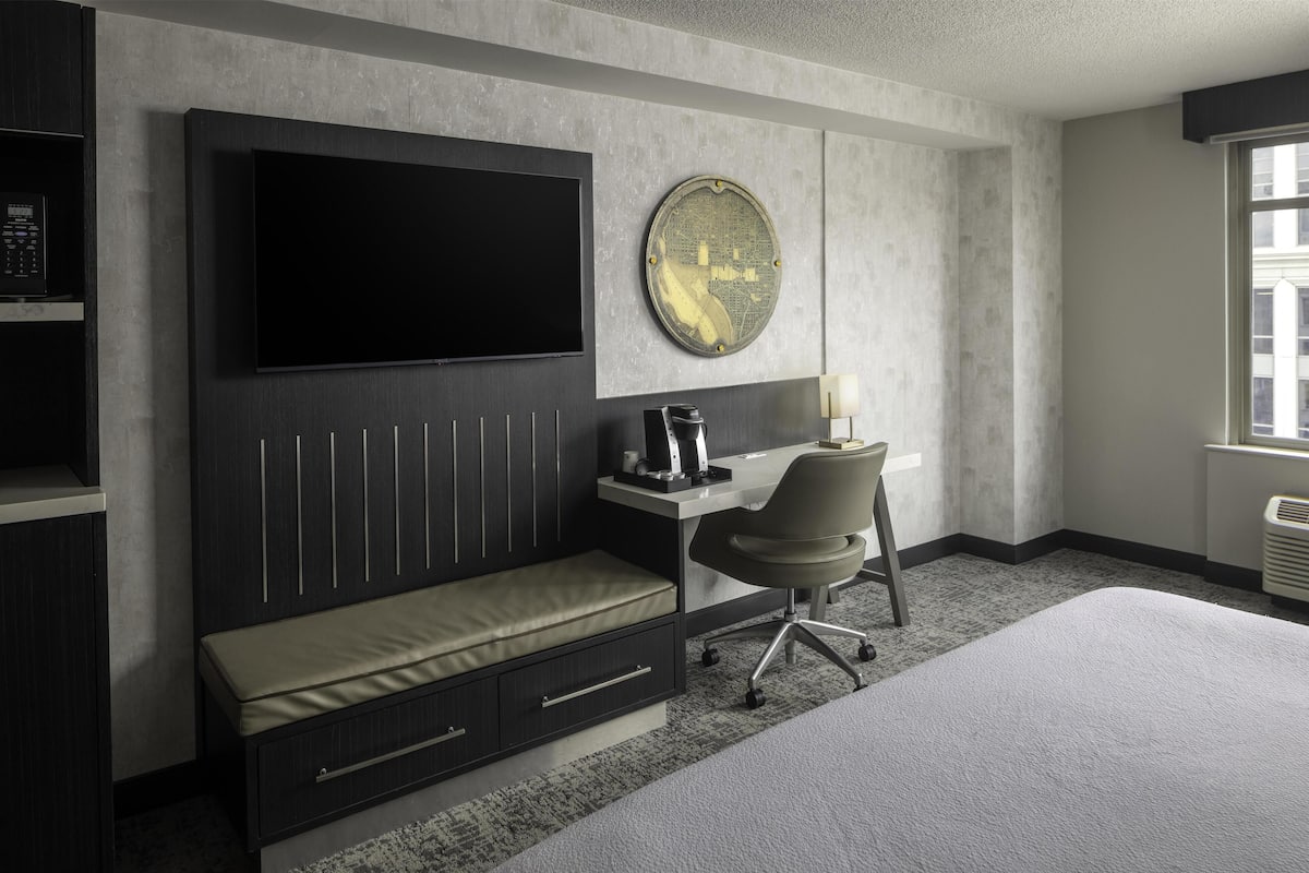 A modern hotel room with a large bed, a desk with a chair, a flat-screen TV, a round decorative wall piece, and a built-in shelf with a coffee maker.